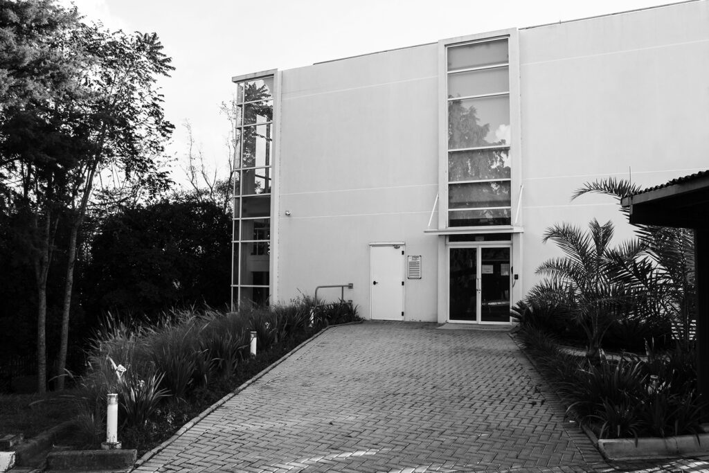Uma foto em preto e branco da entrada de um prédio baixo e branco com calçada e canteiros com plantas.